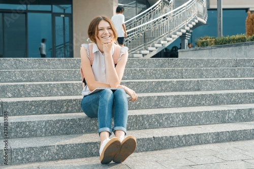 Outdoor portrait of young beautiful smiling student girl with backpack, sitting on the steps in the city