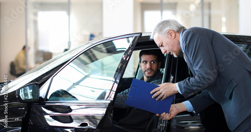 Young man examining a new car in a showroom while the car dealer is explaining its features