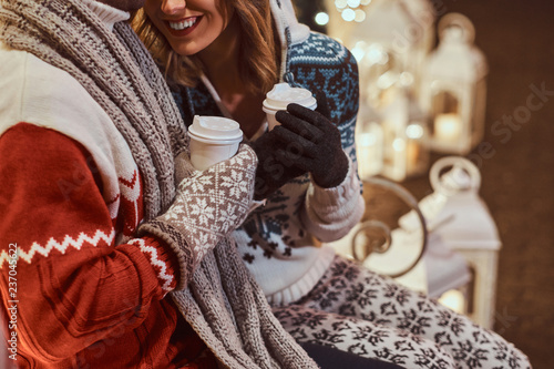 Cropped image of a young romantic couple sitting on a bench and warming with hot coffee in evening street decorated with beautiful lights at Christmas time.