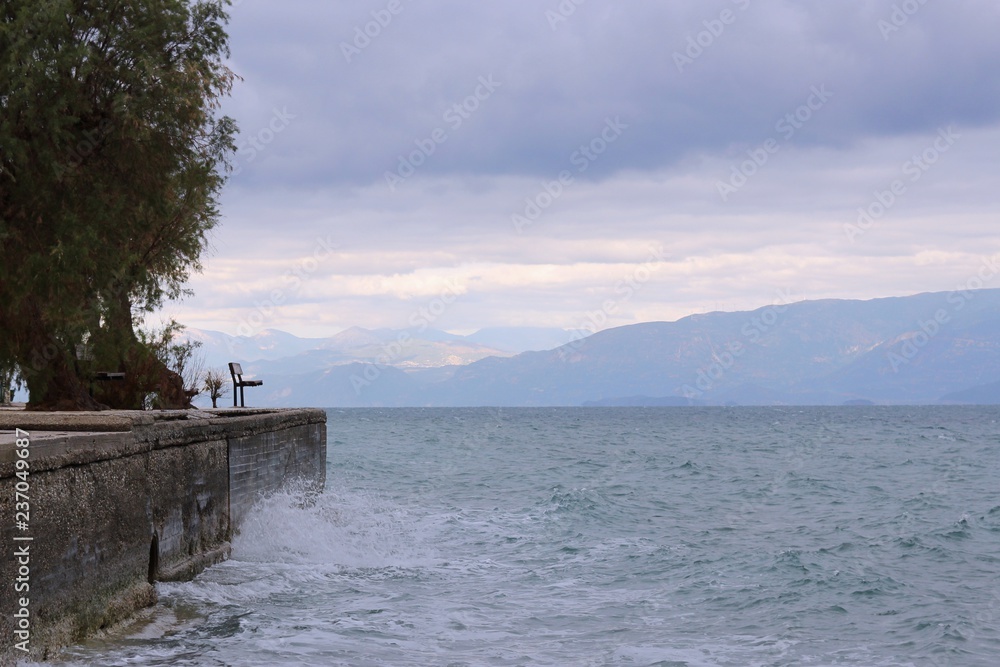 Bench at a dock at stormy weather and big waves at sea clouds and mountains at the background