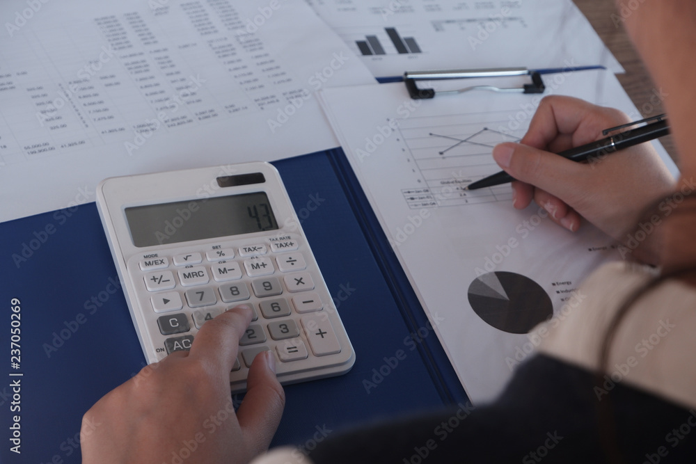 Female working in office, studying using calculator and writing something with documents and chart on table Female working in office, studying using calculator and writing something with documents and chart on table