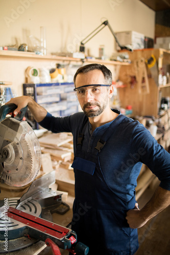 Waist up portrait of mature carpenter posing in workshop  standing next to electric disk saw