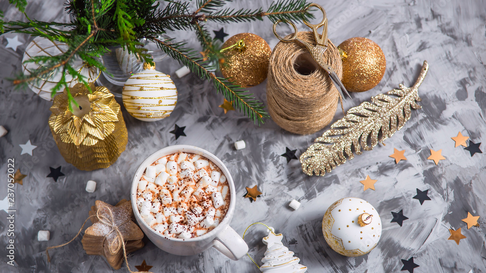 A large cup of cocoa with marshmallow sprinkled with cocoa powder stands on a gray table among Christmas decorations, fir branches, ginger cookies and shiny stars. Top view A large cup of cocoa with marshmallow sprinkled with cocoa powder stands on a gray table among Christmas decorations, fir branches, ginger cookies and shiny stars. Top view