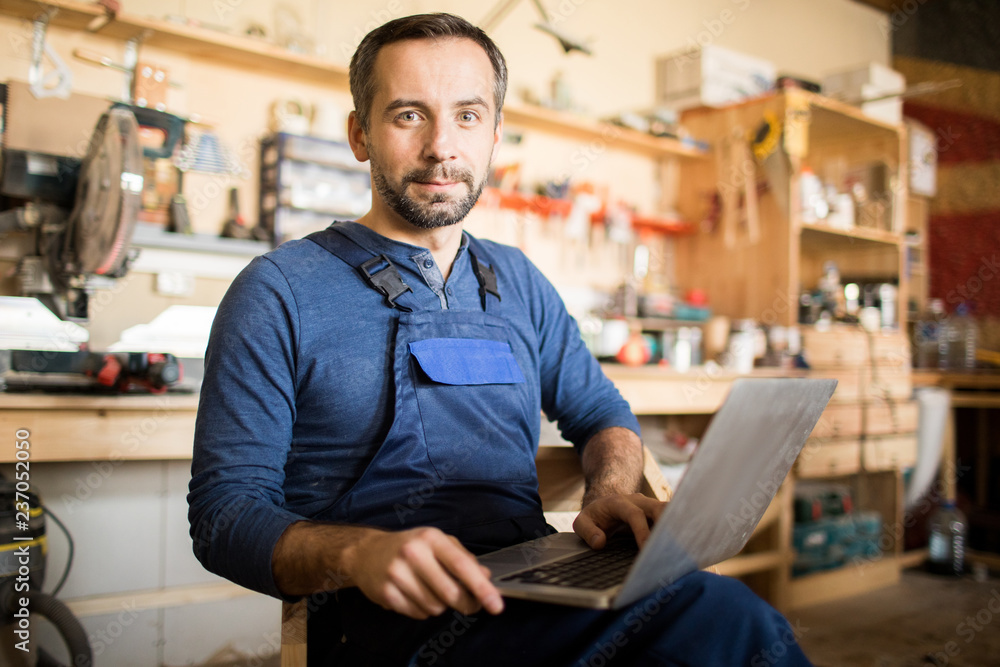 Portrait of mature worker using laptop and looking at camera in workshop interior, copy space Portrait of mature worker using laptop and looking at camera in workshop interior, copy space