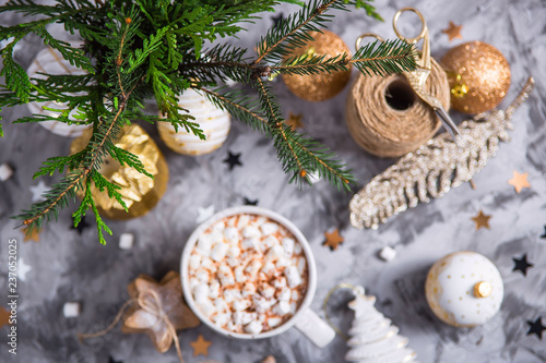 A large cup of cocoa with marshmallow sprinkled with cocoa powder stands on a gray table among Christmas decorations, fir branches, ginger cookies and shiny stars. Top view
