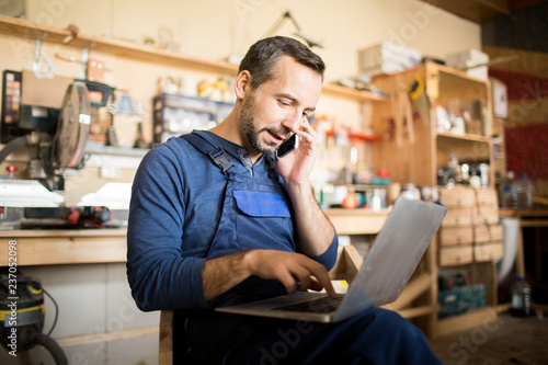Portrait of mature worker using laptop and speaking by smartphone  in workshop interior, copy space