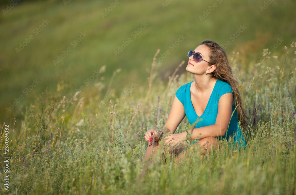 Young and beautiful caucasian girl with long hair in sunglasses sits in the meadow on a sunny day. Pretty woman is sitting on the grass and resting. Young and beautiful caucasian girl with long hair in sunglasses sits in the meadow on a sunny day. Pretty woman is sitting on the grass and resting.