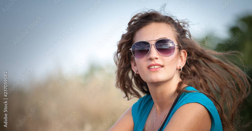 Young and beautiful caucasian girl with long hair in sunglasses sits in the meadow on a sunny day. Pretty woman is sitting on the grass and resting. Young and beautiful caucasian girl with long hair in sunglasses sits in the meadow on a sunny day. Pretty woman is sitting on the grass and resting.