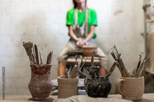 Master class concept.Photo of tools for ceramics work stand against blurred workmanship artisan artist lady in her apron work wear she sit indoor bright workspace