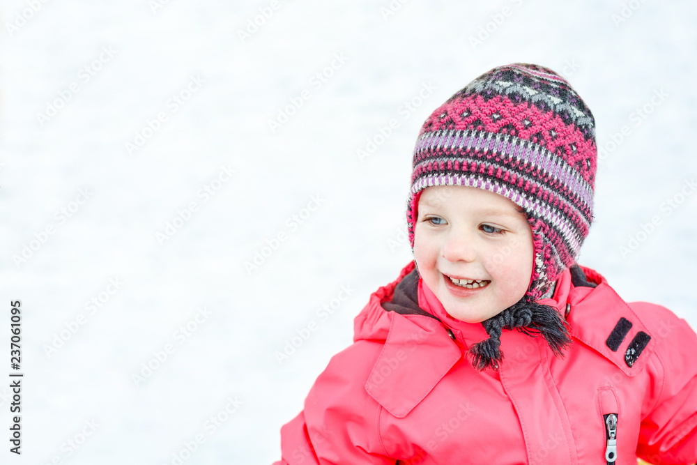 A pretty white girl in a knitted winter hat and pink jumpsuit, smiling and laughing in the snow . Portrait close-up.