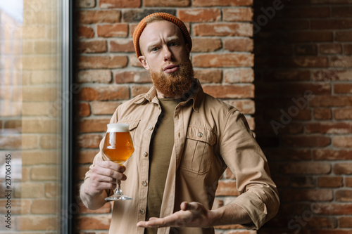 Bearded hipster man with funny emotional face holding glass of beer, drinking alcohol in pub. Positive stylish guy celebrate St. Patrick's Day with drinks photo