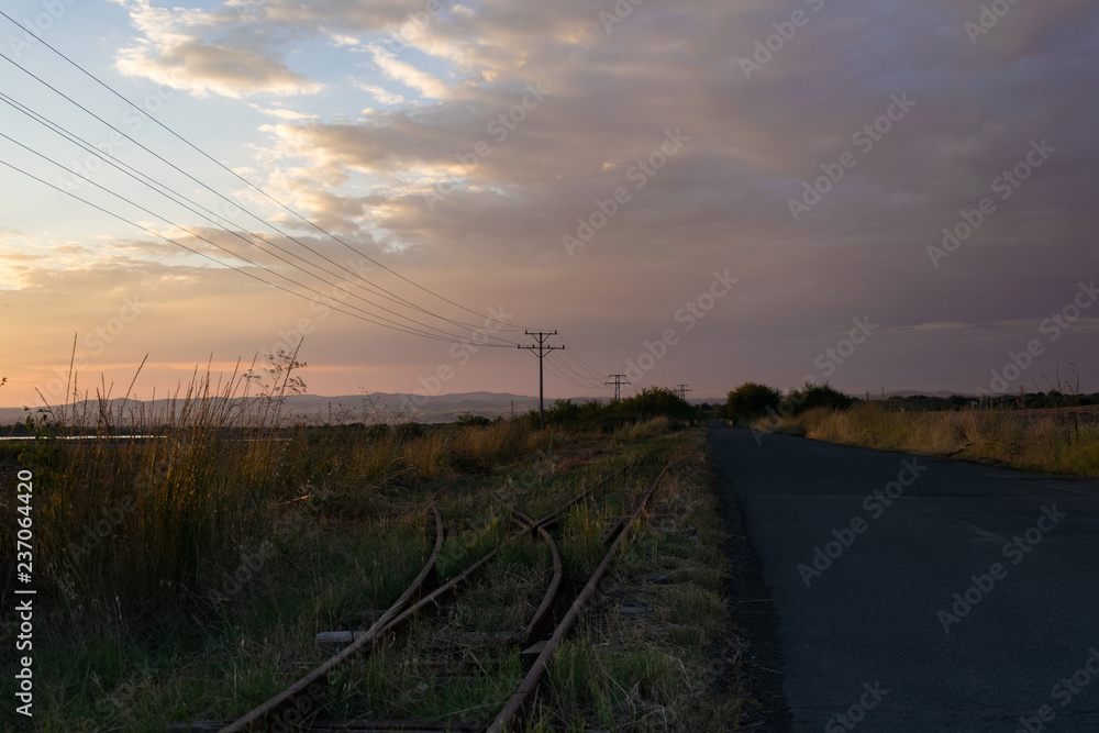 Obraz premium An asphalt road, a train line and power line with and power pylons through the field.