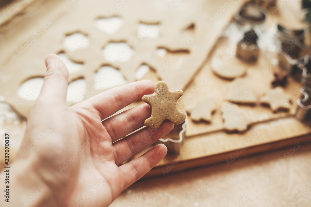 Fototapeta premium Hand holding christmas gingerbread cookie man on background of wooden rolling pin, cookies, metal cutters, christmas decorations on rustic table. Process of making cookies with child