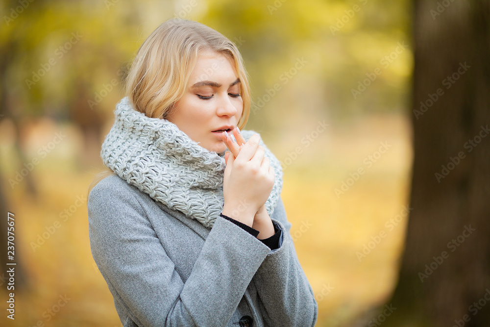 Cold and flu. Young woman in a gray coat walking in the autumn park and warms frozen hand Cold and flu. Young woman in a gray coat walking in the autumn park and warms frozen hand