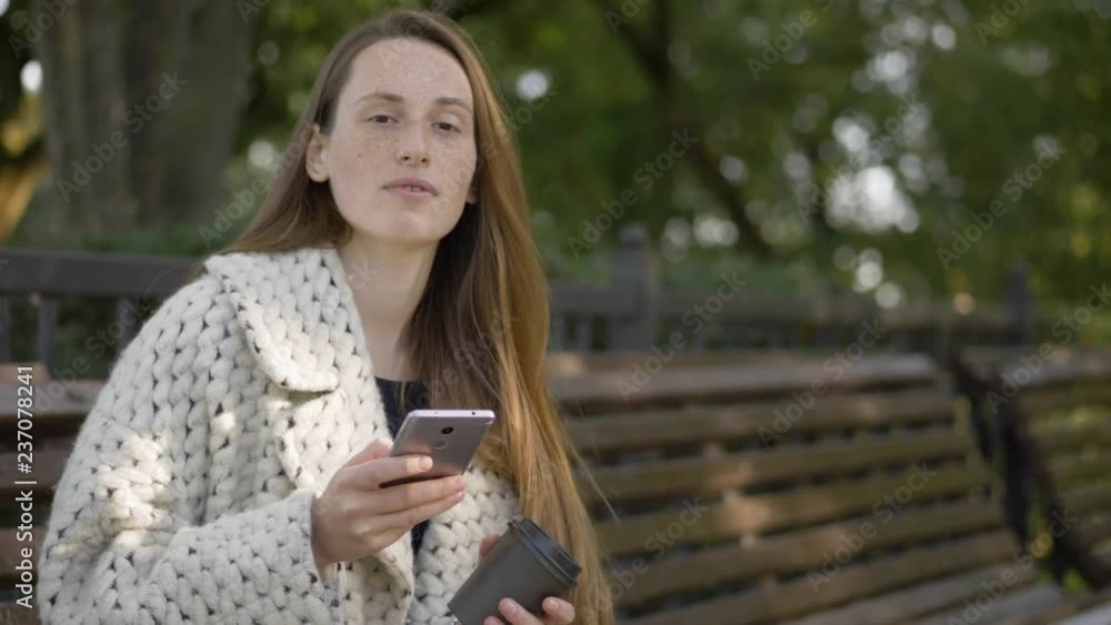 Slow motion portrait of beautiful young woman with freckles, trendy coat using smartphone, checking social networks, enjoying warm evening sitting in city park . Slow motion portrait of beautiful young woman with freckles, trendy coat using smartphone, checking social networks, enjoying warm evening sitting in city park .