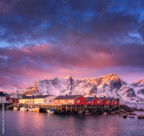 Beautiful fishing village with boats at sunrise  Lofoten islands  Norway. Winter landscape with houses  snowy mountains  sea  boats  colorful sky with clouds. Norwegian traditional red rorbu and rocks