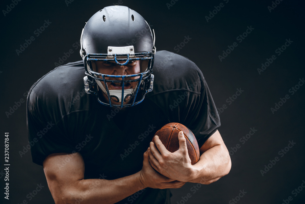 American football caucasian player wearing black uniform with protection holding ball isolated over black studio wall