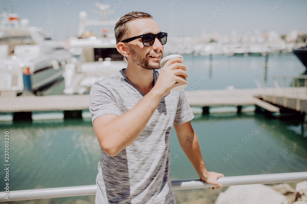 Young man in sunglasses drink coffee to go on summer street with yacht harbor on background. Travel concept