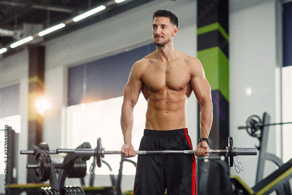 Naklejka premium Attractive muscular guy doing biceps exercises with barbell at the modern gym. Sporty and healthy concept.