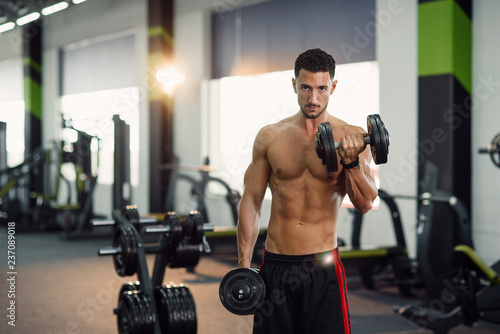 Attractive fitness man with naked torso working out with dumbbells during training biceps in the gym. Sporty and healthy concept.