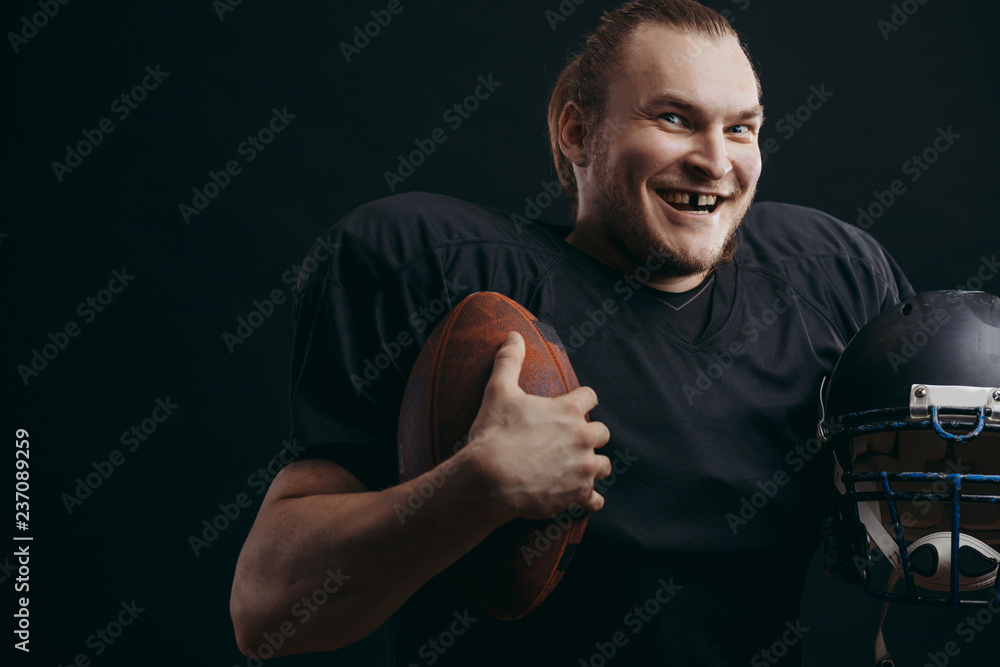 Dramatic portrait of american football player smiling, showing broken front tooth holding his helmet and ball in arms background