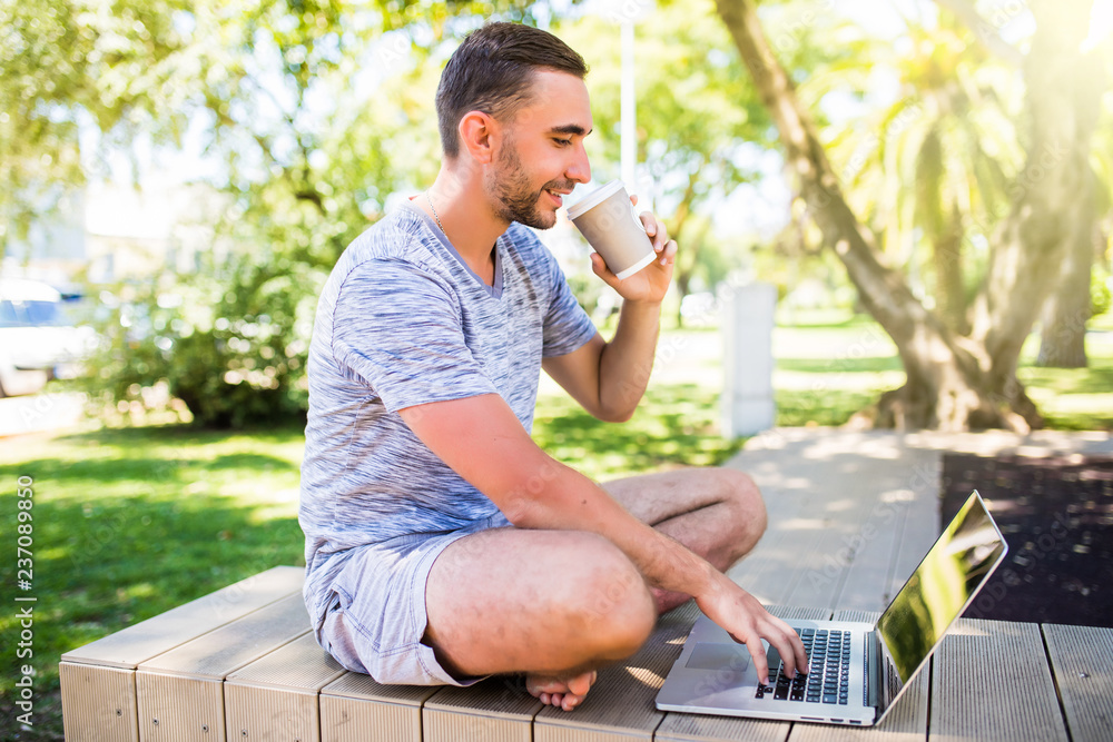 Portrait of young happy man with laptop and cup of coffee on bench in park