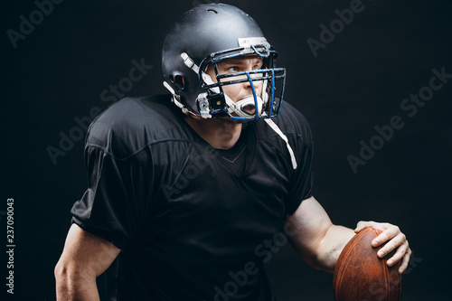People, achievement and sport concept. Athletic american football player in black helmet and jersey posing with a ball isolated on black background