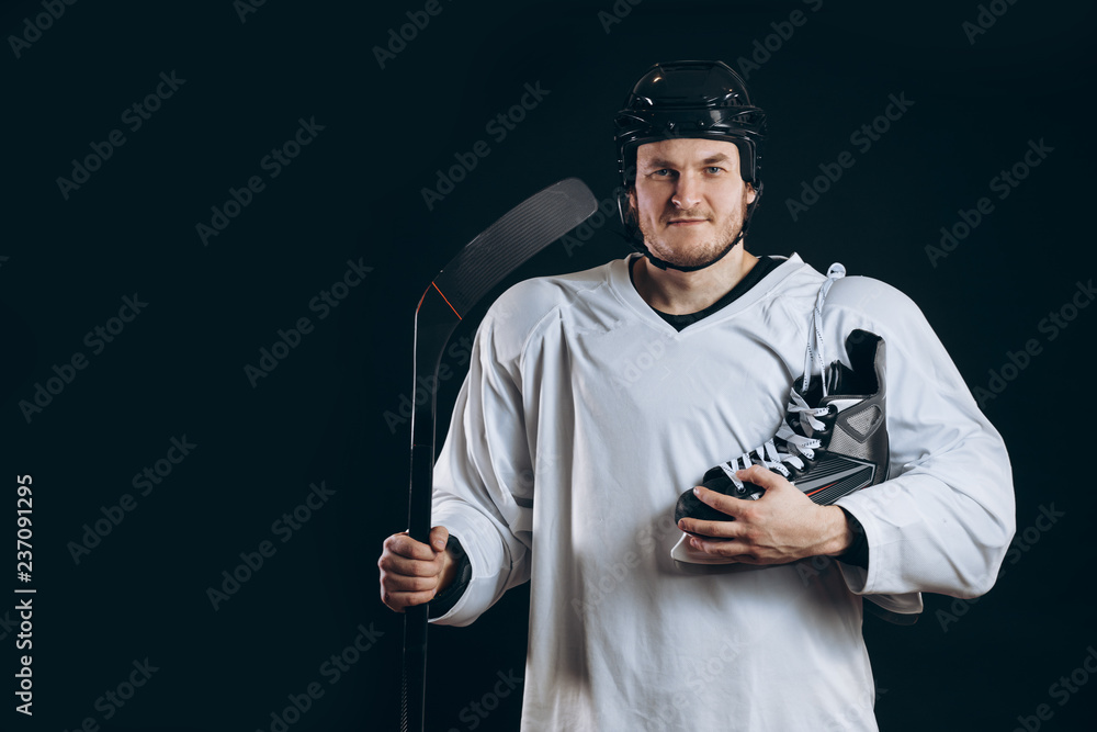 Portrait of a professional hockey player in a protective sportswear and helmet posing with hockey stick and one skate in hands on a black background. Portrait of a professional hockey player in a protective sportswear and helmet posing with hockey stick and one skate in hands on a black background.