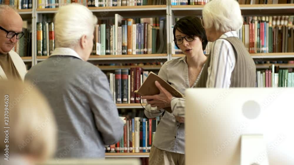 Young female teacher in glasses communicating with three elderly people and answering their questions during lesson in library Young female teacher in glasses communicating with three elderly people and answering their questions during lesson in library