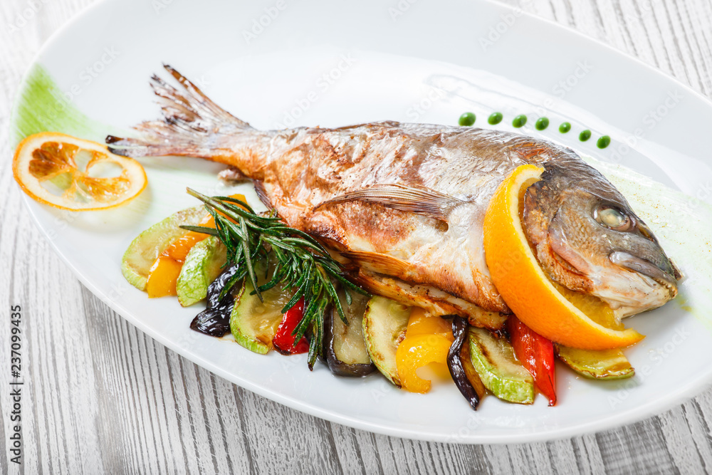 Grilled dorado fish with baked vegetables and rosemary on plate on wooden background close up. Healthy food. Top view Grilled dorado fish with baked vegetables and rosemary on plate on wooden background close up. Healthy food. Top view