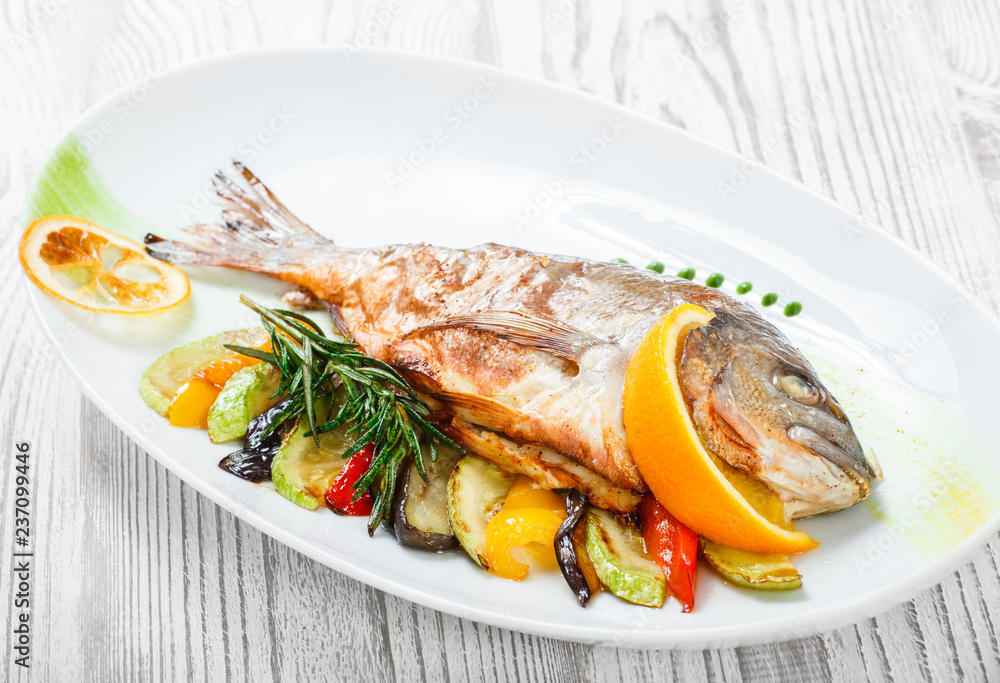 Grilled dorado fish with baked vegetables and rosemary on plate on wooden background close up. Healthy food. Top view Grilled dorado fish with baked vegetables and rosemary on plate on wooden background close up. Healthy food. Top view