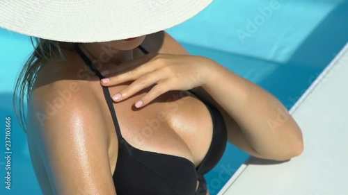 Rich female wearing big white hat stroking body in swimming pool, sunbathing