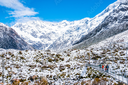 Group of people across the river by a swing bridge in Hooker Valley Track covered with white snow after a snowy day. Mount Cook National Park.
