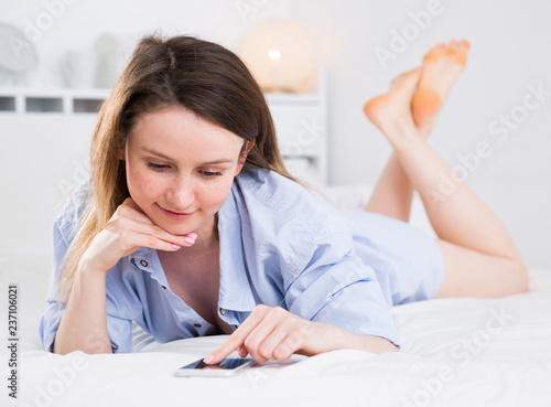 Smiling young woman  in blue shirt relaxing in bed with smartphone