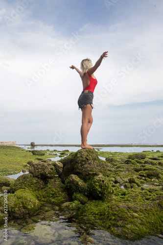 Young woman in a red swimsuiti with raised hands at the beach, standing on the moss covered rocks, full-length portrait with natural sunlight, wellbeing holiday concept