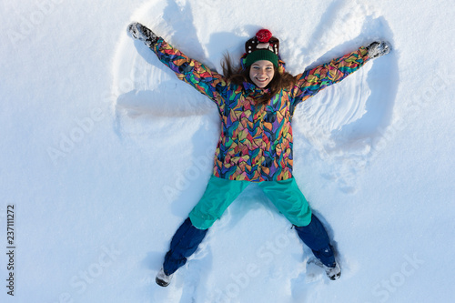 High angle view of happy girl lying on snow and moving her arms and legs up and down creating a snow angel figure. Smiling woman lying on snow in winter holiday with copy space