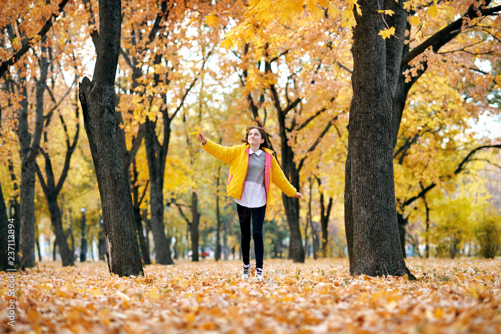 Happy teen girl is running in autumn park with big maple's leaf. Bright yellow leaves and trees.