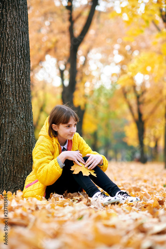 Sad teen girl sits near tree in autumn park. Bright yellow leaves and trees.