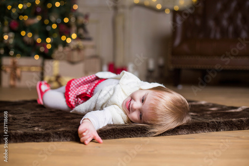 small female child is lying on the floor against the lights of the Christmas tree and smiling funnyly photo