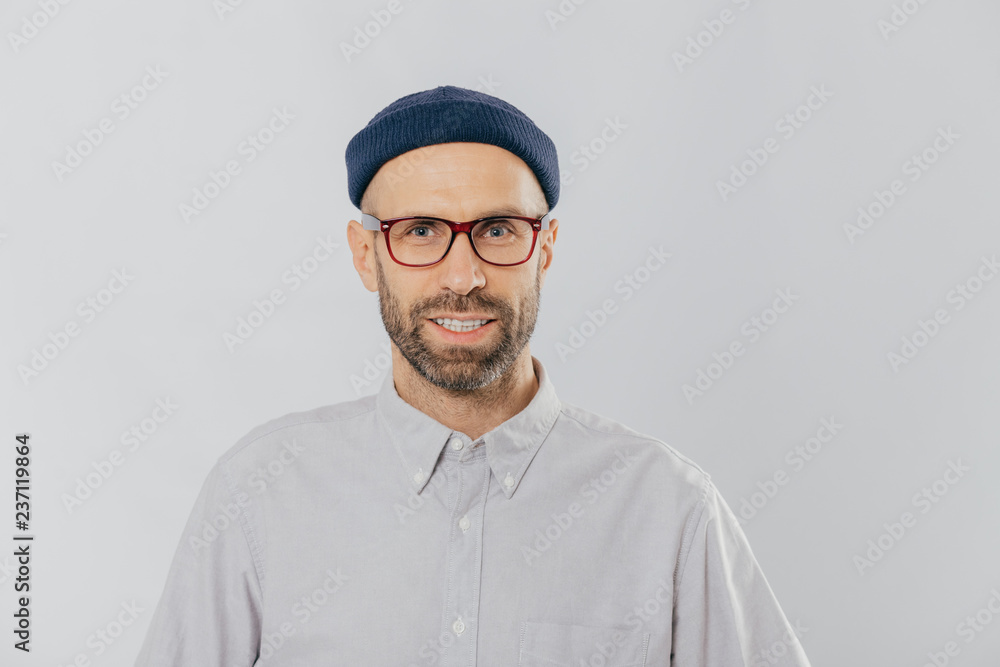 Horizontal view of pleased smiling man wears glasses, hat and shirt, being satisfied with good news, models against white background. Unshaven hipster rejoices new purchase. Fashion concept. Horizontal view of pleased smiling man wears glasses, hat and shirt, being satisfied with good news, models against white background. Unshaven hipster rejoices new purchase. Fashion concept.