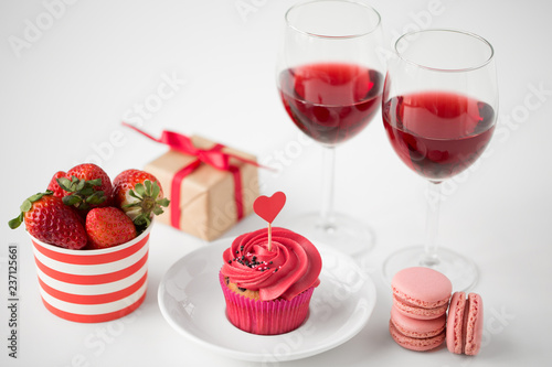 valentines day and sweets concept - close up of frosted cupcake with red heart shaped cocktail stick, macarons, strawberries, glasses of wine and gift box on white background