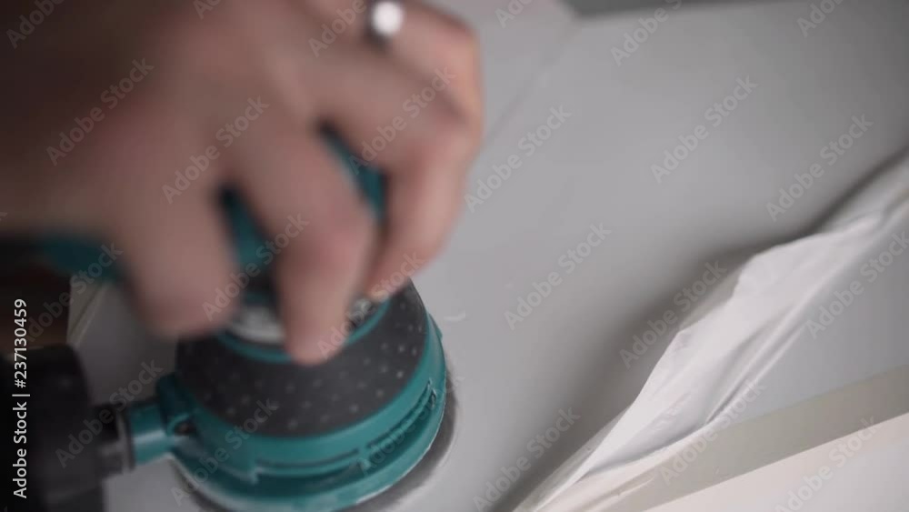 Man's hand is holding green round-shaped electronic polisher and working with it on white surface in light room. Man's hand is holding green round-shaped electronic polisher and working with it on white surface in light room.