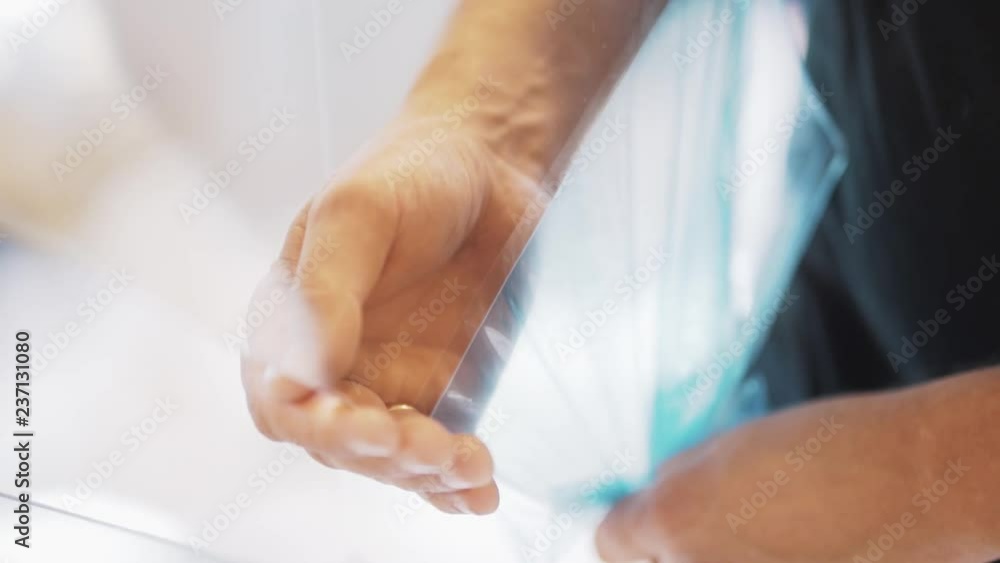 Mature man dressed in in black t-shirt with hands is carefully removing blue film from surface of large square shaped glass in light studio. Mature man dressed in in black t-shirt with hands is carefully removing blue film from surface of large square shaped glass in light studio.