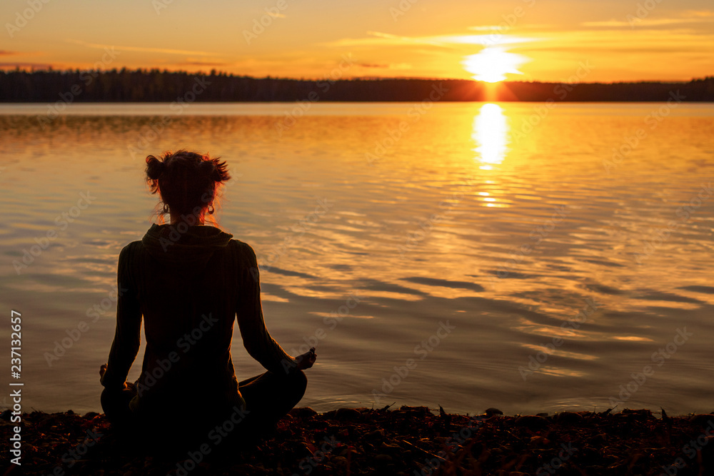 Silhouette of young woman practising yoga outdoor. Meditation on the shore of the lake during sunset. Fitness and healthy lifestyle concept.