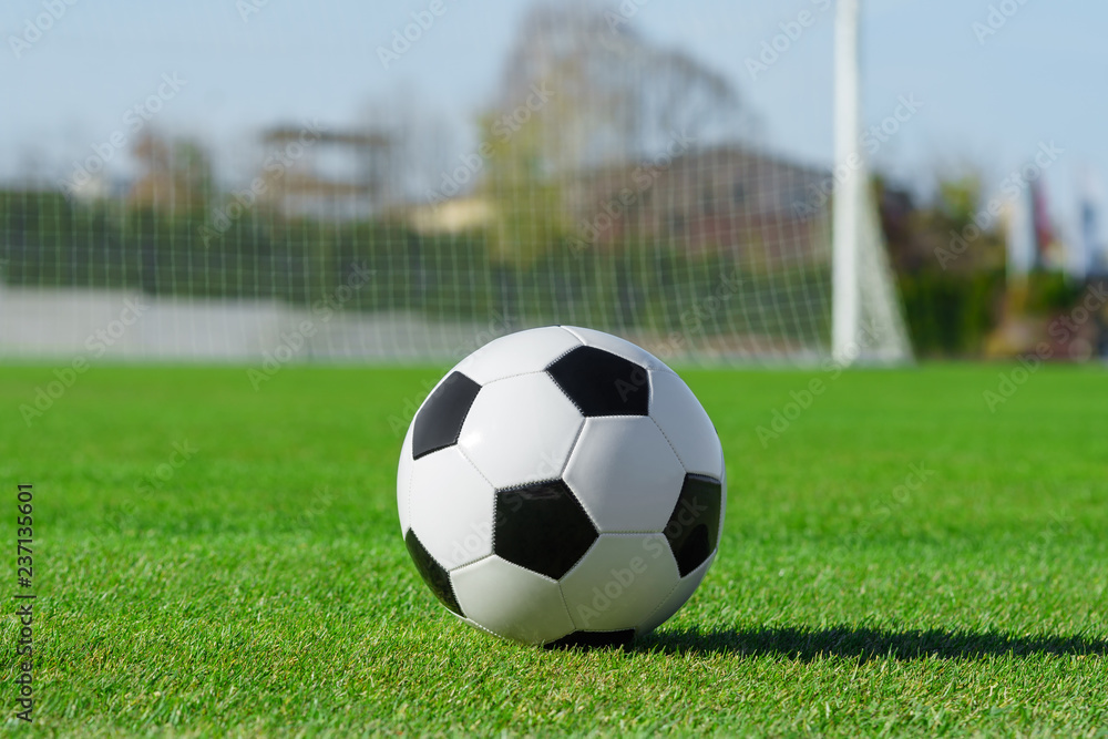 Classic soccer ball lies on the bright green grass on the football field against the background of the stands for fans and the football goal at a sports stadium close-up in a large sports center