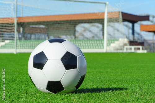 Classic soccer ball lies on the bright green grass on the football field against the background of the stands for fans and the football goal at a sports stadium close-up in a large sports center