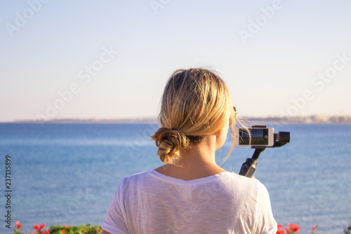 Video blogger girl. Operator with action camera on stabilized grip with gimbal. Happy young woman shoots 4k footage on event outdoor