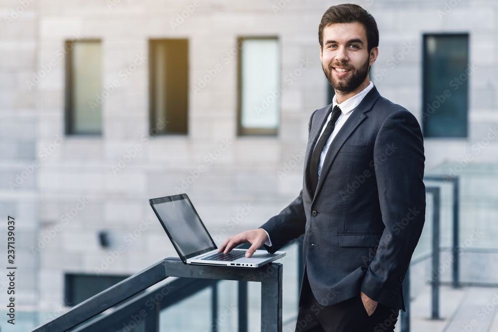 Cheerful businessman in a suit is smiling widely. Stands in the background of the office with a laptop Cheerful businessman in a suit is smiling widely. Stands in the background of the office with a laptop