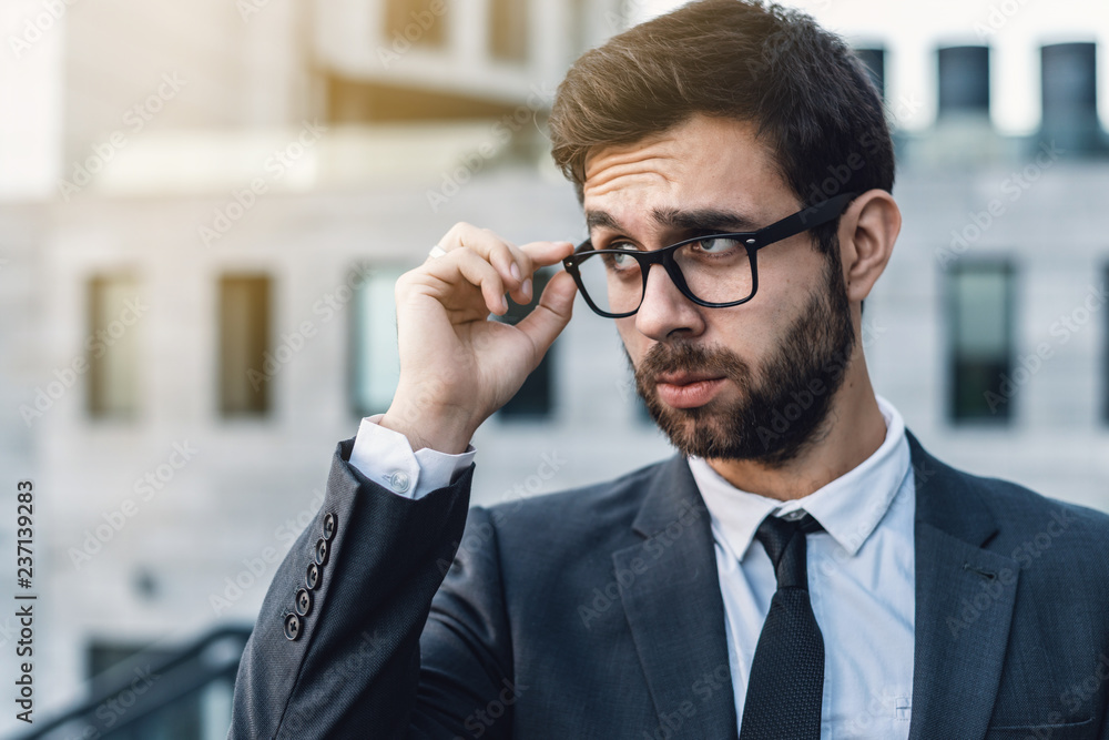 Portrait of a businessman is touching eyeglasses in a classic suit against an office building. Looking from under the glasses to the side touching the glasses. Portrait of a businessman is touching eyeglasses in a classic suit against an office building. Looking from under the glasses to the side touching the glasses.