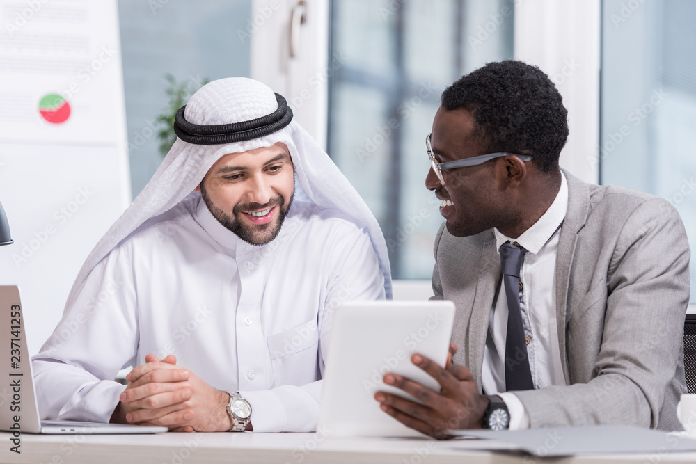 Fototapeta premium Multiethnic partners looking on digital tablet and smiling in modern office